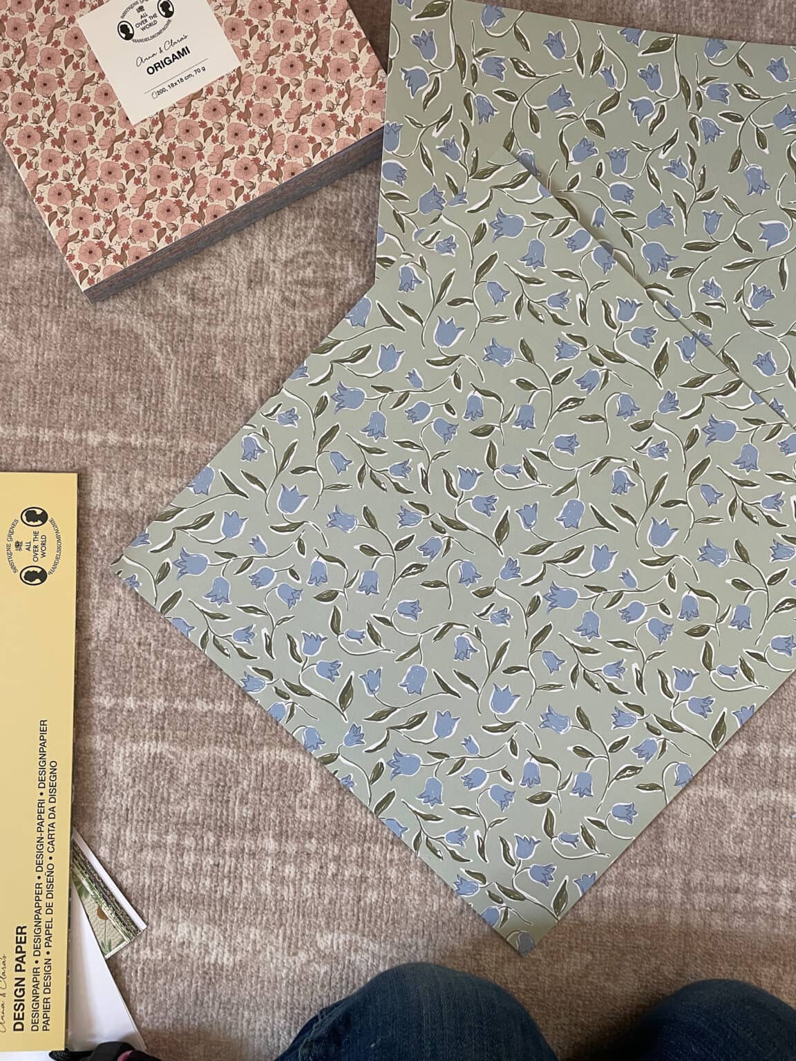 A person sits on a beige carpet with floral craft paper in blue and green tones, a pink floral box, and a yellow pack labelled design paper arranged around them. Only their knees and part of their legs are visible.