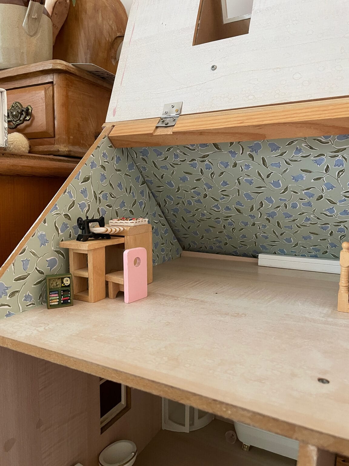 A miniature room in a doll’s house with floral wallpaper, featuring a small sewing machine on a wooden table, a pink iron, and a green sewing kit on the floor.