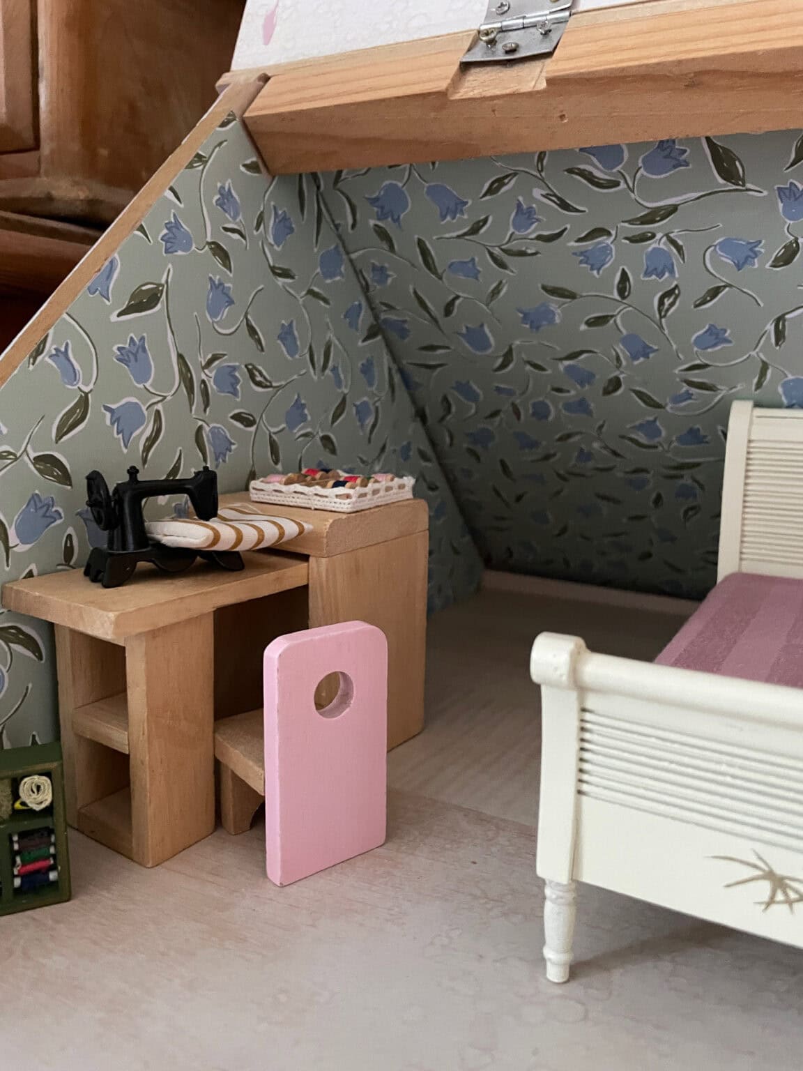 A miniature room with a sloped ceiling, featuring a small wooden desk with a sewing machine and fabric, a pink chair, a floral-patterned wallpaper, and part of a white bed with pink bedlinen.