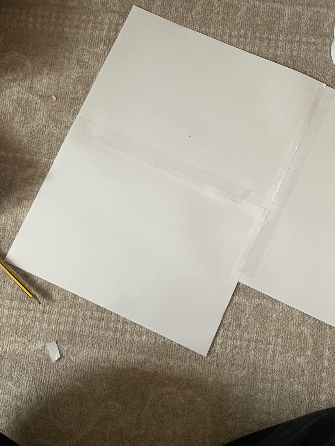 A large white sheet of paper taped at the seams sits on a beige carpet. A pencil and a small piece of tape are nearby. Part of a black object appears at the bottom edge of the image.