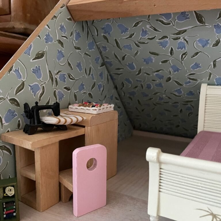 A miniature sewing room with a small black sewing machine on a wooden desk, a pink stool, an ironing board, and a bed. The slanted wall has blue floral wallpaper.