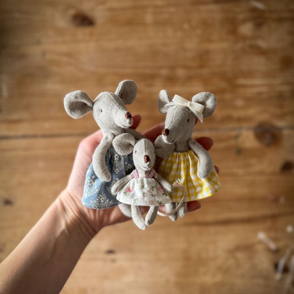 A hand holds three small, fabric toy mice—made using a mouse sewing pattern—each dressed in patterned dresses: one blue, one yellow check with a bow, and a smaller pink floral, all against a wooden background.