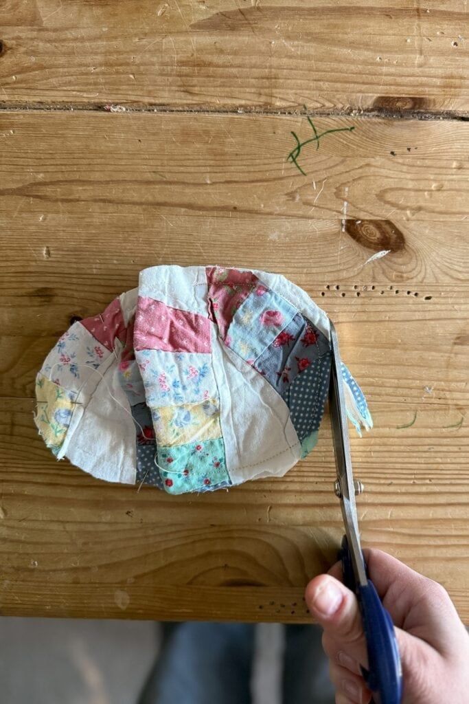A hand holding scissors cuts the edge of a colourful patchwork fabric piece on a wooden table. The fabric, adorned with floral and patterned patches, includes whimsical shapes like fabric eggs sewn into the design.