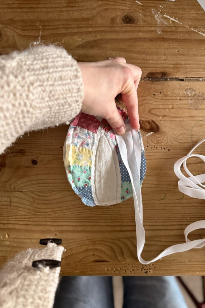 A hand holds a round, patchwork fabric egg with colourful patterns on a wooden table, about to tie a white ribbon round it. Part of a cream-coloured jumper sleeve and blue jeans are visible.