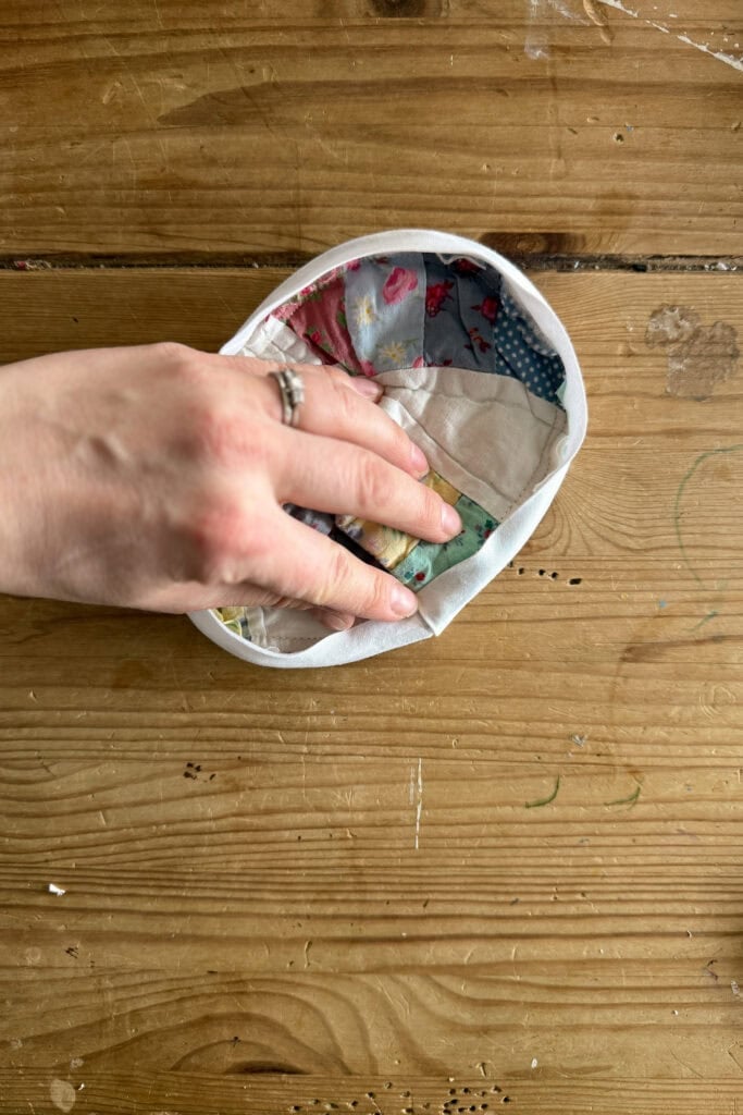 A hand with rings holds a colourful, patterned fabric bowl or pouch filled with fabric eggs on a wooden surface, with light and dark wood grain and small green marks visible on the table.