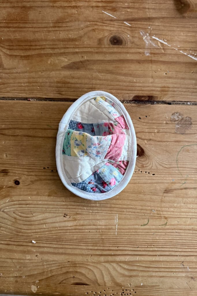 A quilted fabric egg, oval in shape with a white border and colourful patchwork design, sits on a wooden surface.