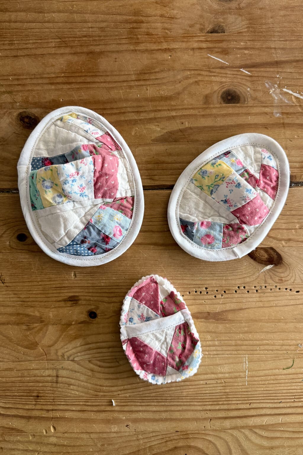 Three quilted fabric eggs with patchwork patterns in pastel colours are arranged on a wooden surface. The two larger fabric eggs sit above a smaller one, each displaying a charming mix of floral and patterned fabrics.
