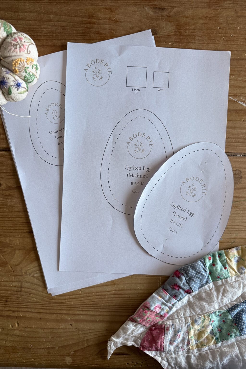Two printed sewing patterns, shaped like eggs, rest on a wooden table surrounded by fabric pieces and a partially visible hand-stitched quilt. The patterns are labelled Quilted Egg (Medium/Small) BACK.