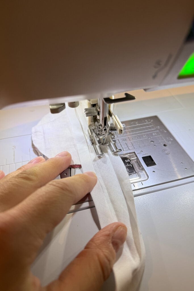 A close-up view of a person's hand guiding white fabric through a sewing machine as they sew a straight seam, possibly creating fabric eggs or other decorative items.