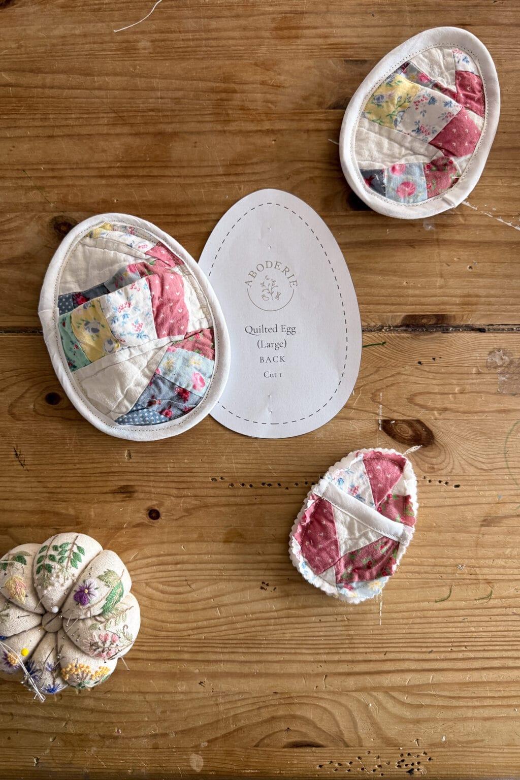 A wooden table with three fabric eggs featuring quilted designs, a fabric pin cushion with floral embroidery, and a paper pattern labelled Broderie Quilted Egg (Large) Back Cut 1.