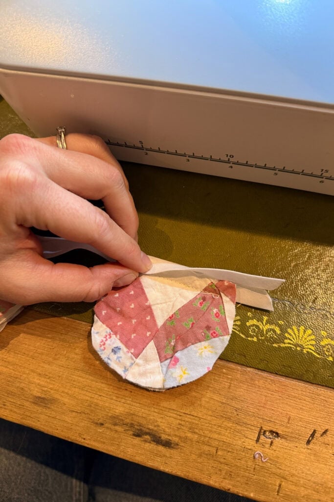 A hand holds a round, patchwork fabric egg with various patterns next to a strip of fabric, on a wooden table beside a sewing machine.