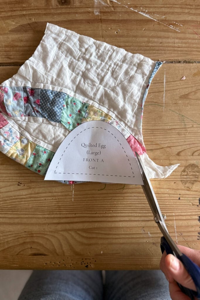 A person uses scissors to cut quilted fabric eggs along the edge of a paper template labelled “Quilted Egg (Large) FRONT A Cut.” The colourful patchwork fabric rests on a wooden table.
