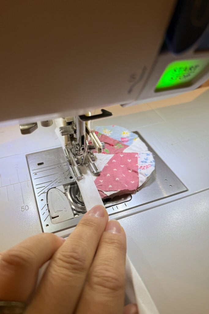 A close-up of a person guiding fabric with a colourful patchwork design—featuring playful fabric eggs—through a sewing machine, stitching a white strip onto the edge. The sewing machine’s needle and presser foot are clearly visible.