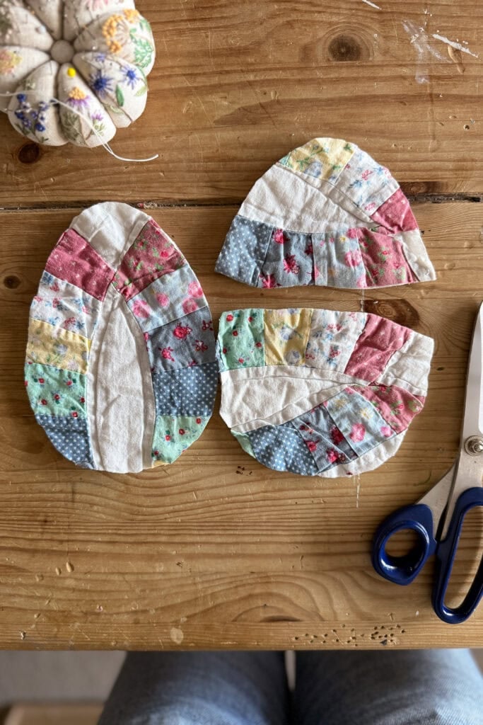 Three quilted fabric eggs with colourful, patterned strips are arranged on a wooden table next to a pincushion and a pair of blue-handled scissors; part of someone’s legs in jeans is visible at the bottom.