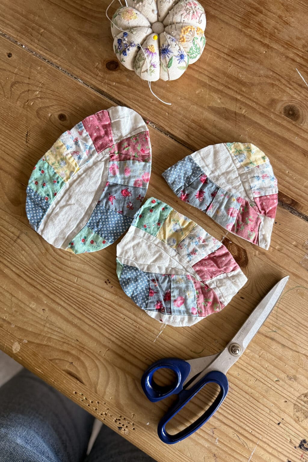 Three colourful, quilted fabric pieces  are laid on a wooden table next to blue-handled scissors and a round, floral-patterned pincushion with pins.