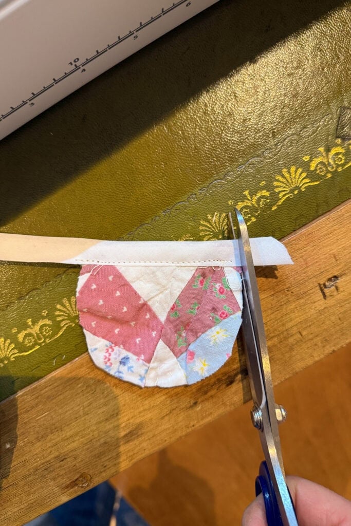 A hand uses scissors to cut a strip of white fabric off a small, quilted patchwork piece with pink, white, blue, and green patterns—perfect for crafting fabric eggs. The scene is on a wooden table with a green, decorated surface nearby.