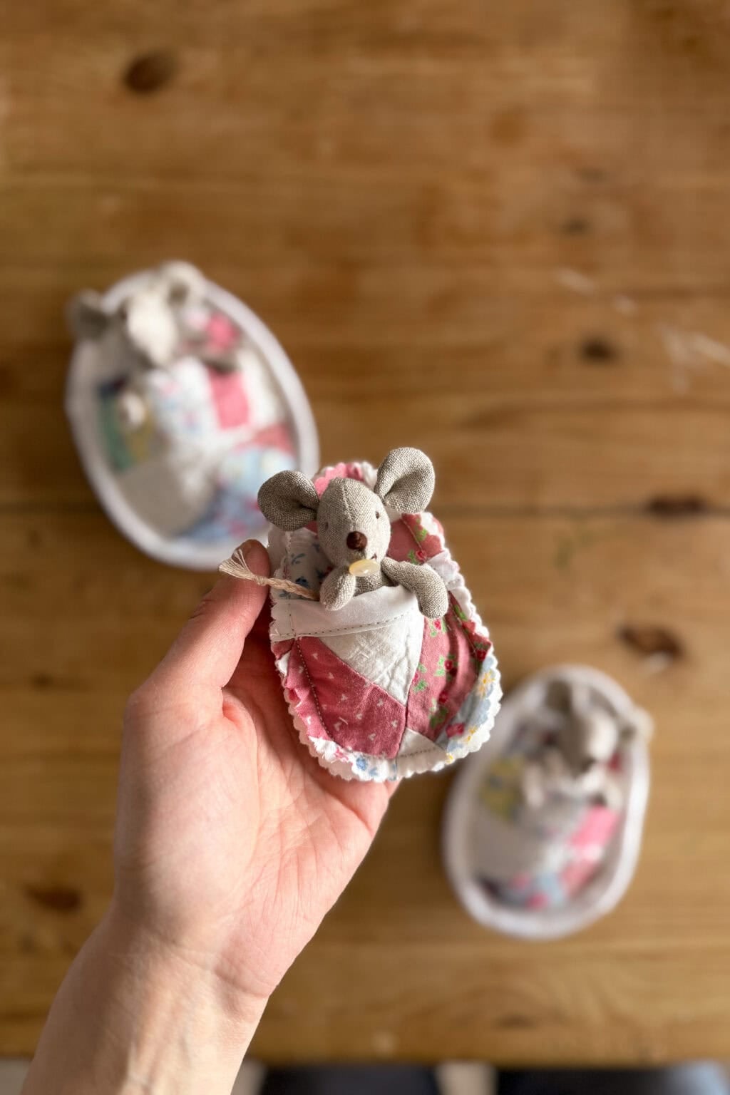 A hand holds a small, stuffed toy mouse wrapped in a quilt. Several fabric eggs rest on a wooden table in the background.