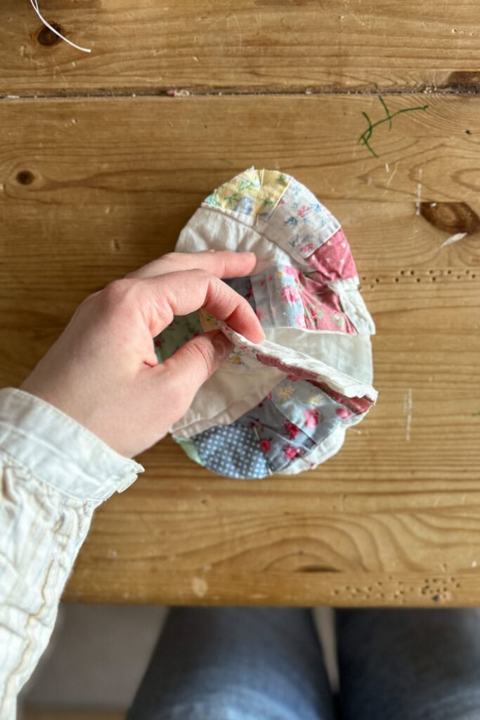 A hand holds a colourful, partially sewn piece of patchwork fabric—perhaps for fabric eggs—on a wooden table, with scattered green thread nearby. The person wears a long-sleeved shirt and jeans.