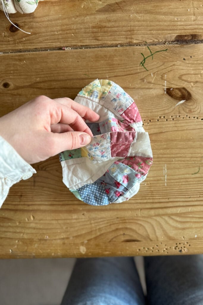 A hand holds a fabric egg, crafted from colourful patchwork with floral and polka dot patterns. This unique piece rests on a wooden surface with visible grain and a few green threads scattered nearby.