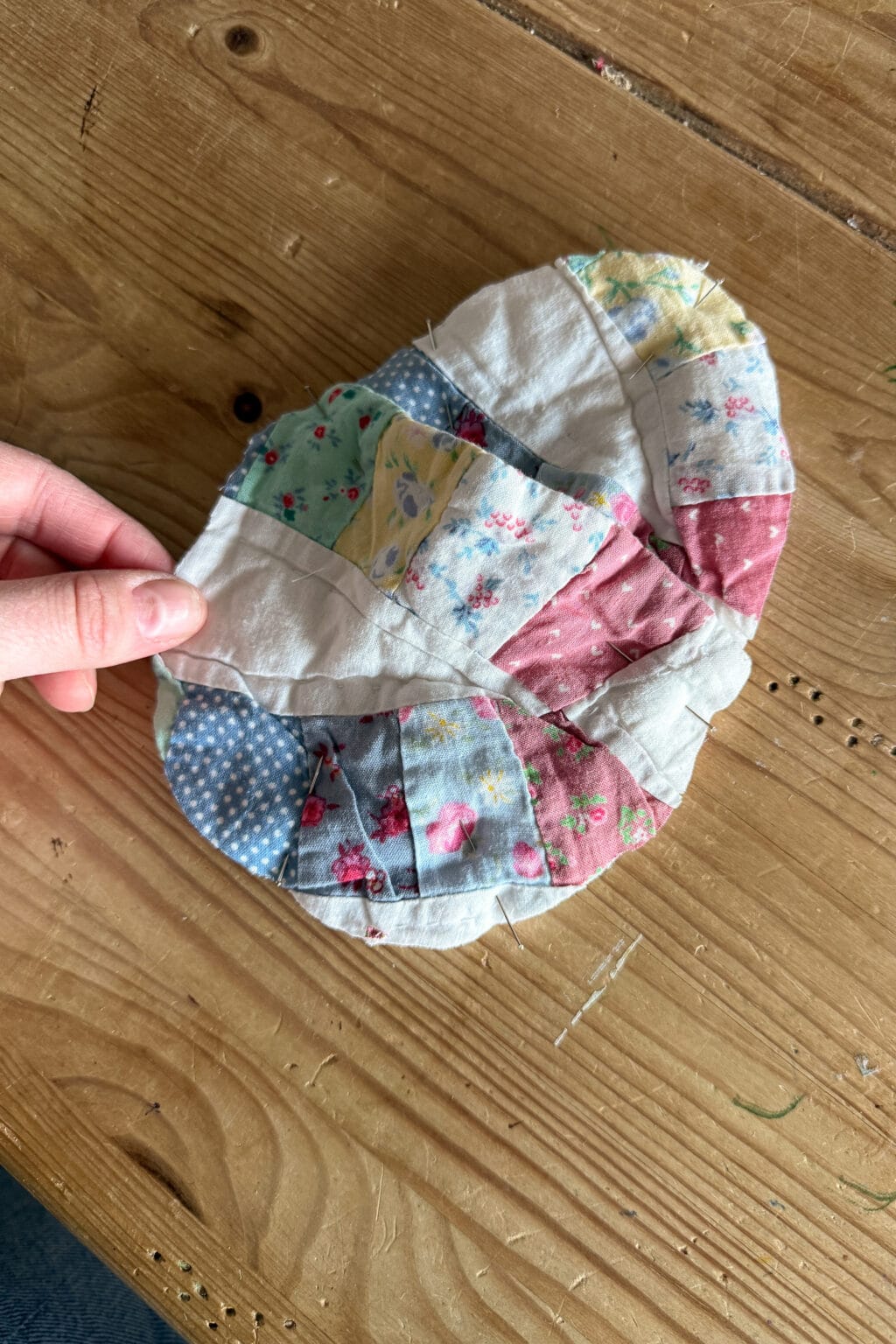 A hand holds a patchwork quilt piece made of curved, colourful fabric segments in floral and patterned prints placed on a wooden table.