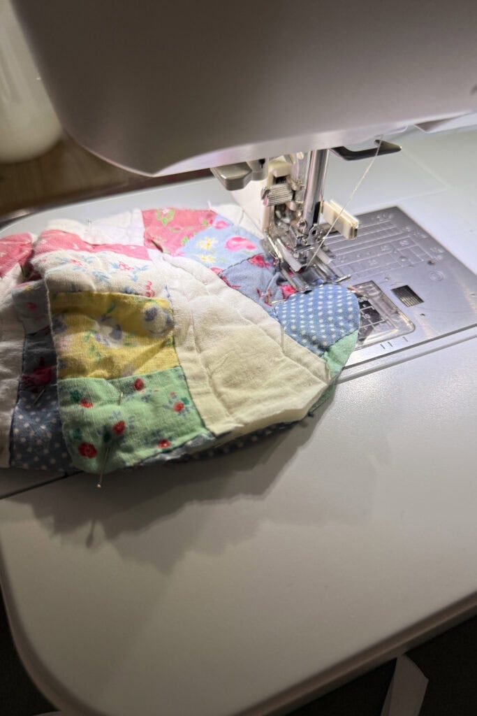 A close-up of a sewing machine stitching a colourful patchwork quilt made of various patterned fabric squares and whimsical fabric eggs. The quilt is partially under the machine's needle and presser foot.