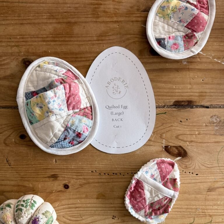 A wooden table with fabric eggs in quilted patchwork patterns, a fabric pincushion, and a paper template labelled Aboderie Quilted Egg (Large) Back Cut 1. The colourful fabric eggs add charm to the sewing scene.