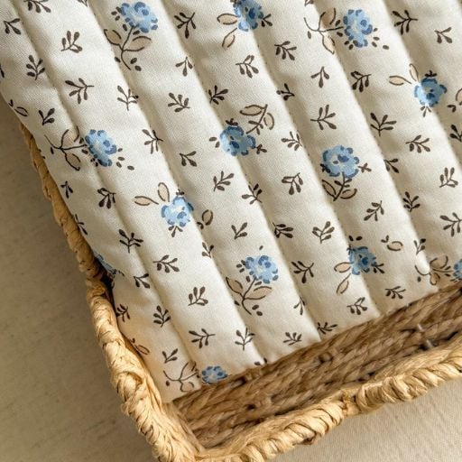 A close-up of a wicker basket lined with quilted white fabric decorated with small blue and beige floral patterns. The lining, enhanced by vertical stitching, holds a few charming fabric eggs that add a whimsical touch.