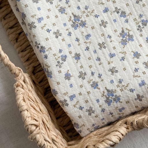 A close-up of folded, textured cream-coloured fabric with a small blue and brown floral print and soft material, lying in a woven basket beside charming fabric eggs. The pattern adds a cosy, delicate appearance.