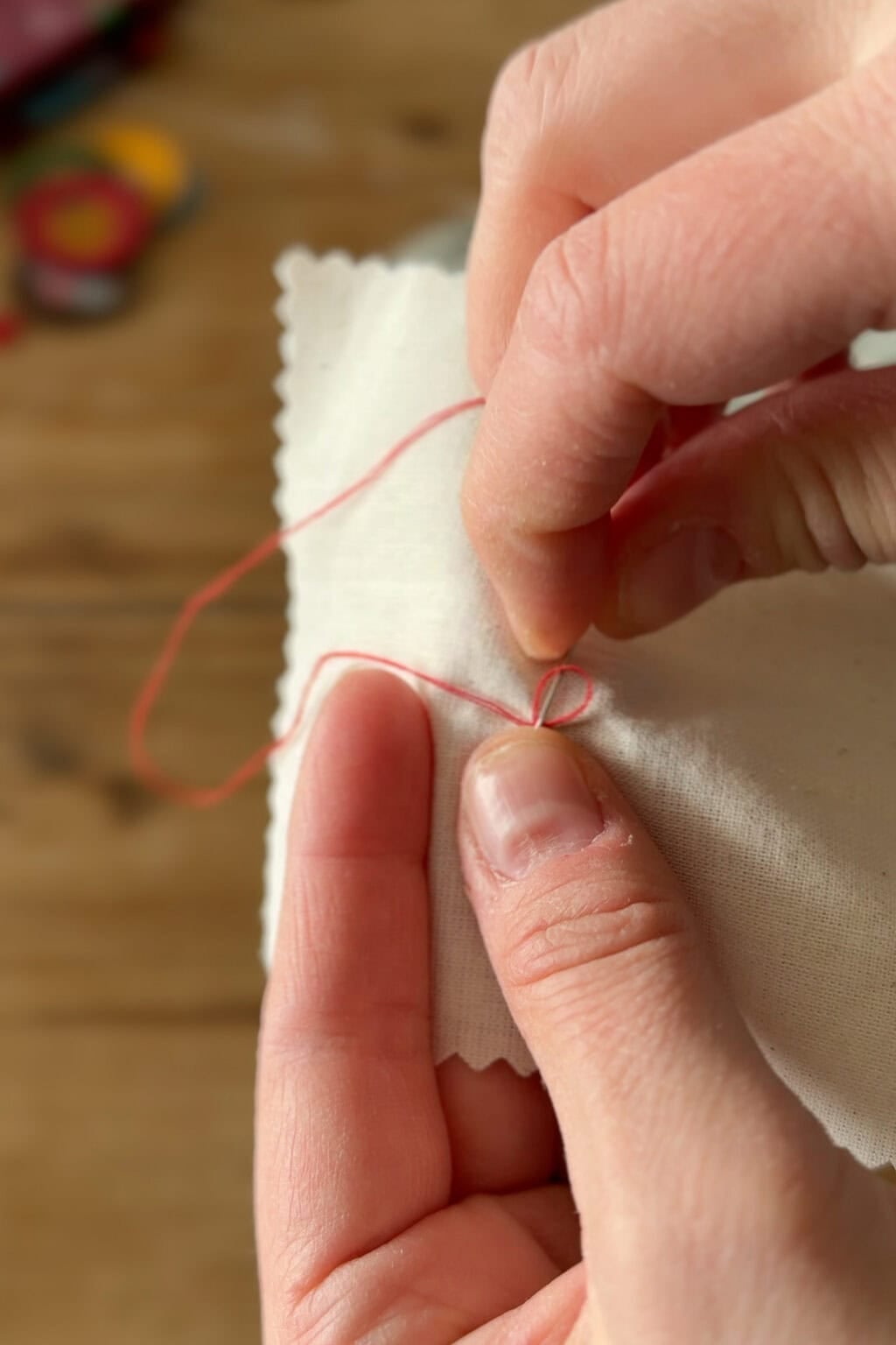 A close-up of two hands sewing a piece of white fabric with red thread, showing the needle being pulled through the material. The wooden surface of a table is blurred in the background.