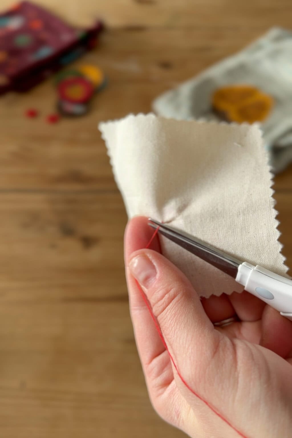 A hand holds a piece of cream-coloured fabric while using a seam ripper to remove red stitches. The background shows blurred sewing supplies on a wooden surface.