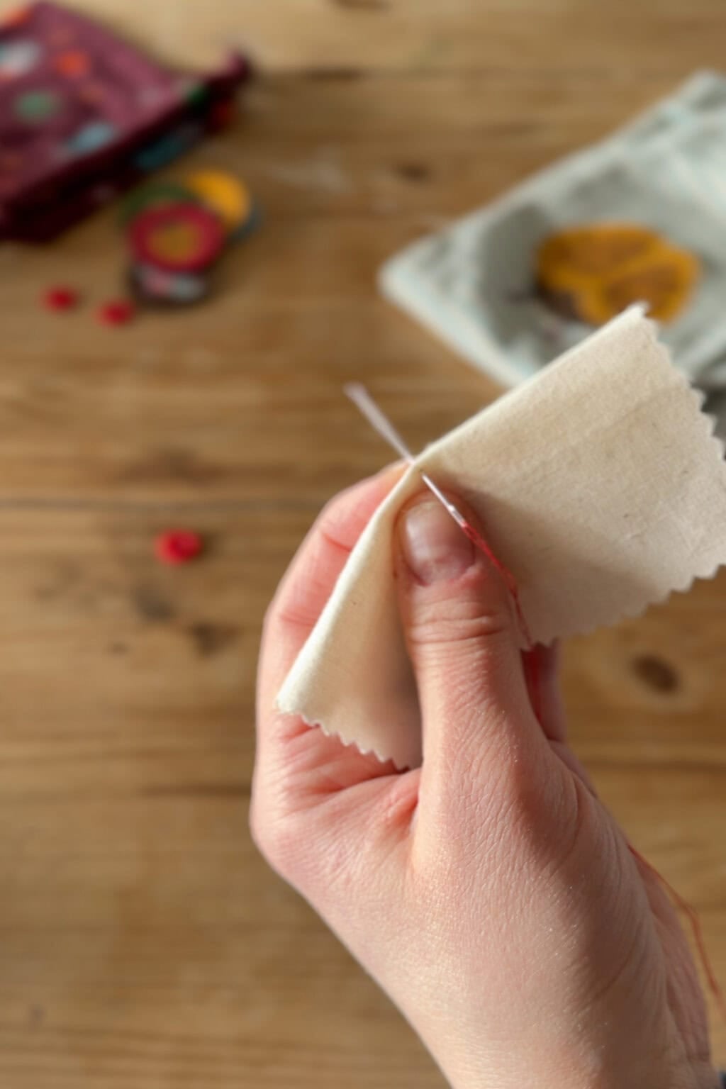 A close-up of a hand sewing two pieces of cream-coloured fabric together with a needle and thread. The background shows a wooden table with blurred sewing supplies.