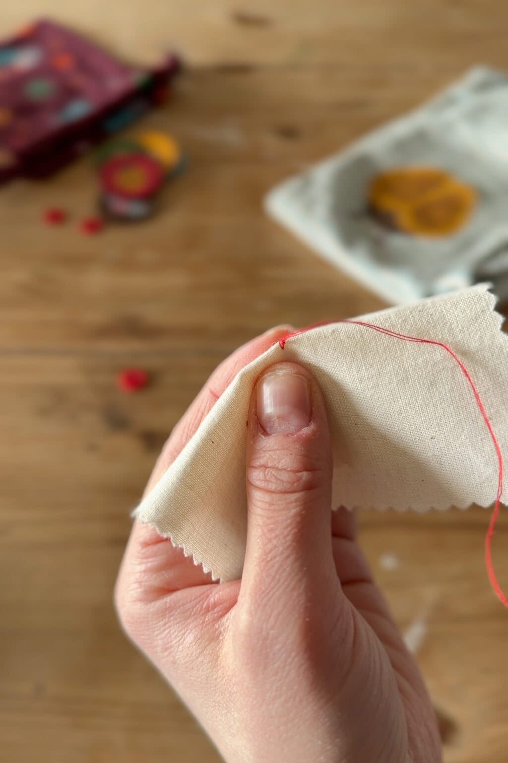 A hand holds a piece of cream-coloured fabric while sewing it with a needle and red thread; a wooden table and sewing materials are visible in the blurred background.