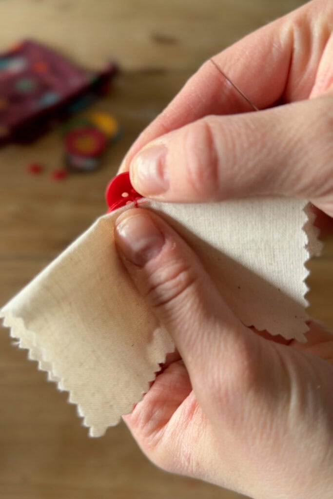 Close-up of hands sewing a red button onto a piece of cream-coloured fabric with pinked edges, using a needle and thread. A blurred background shows sewing supplies.