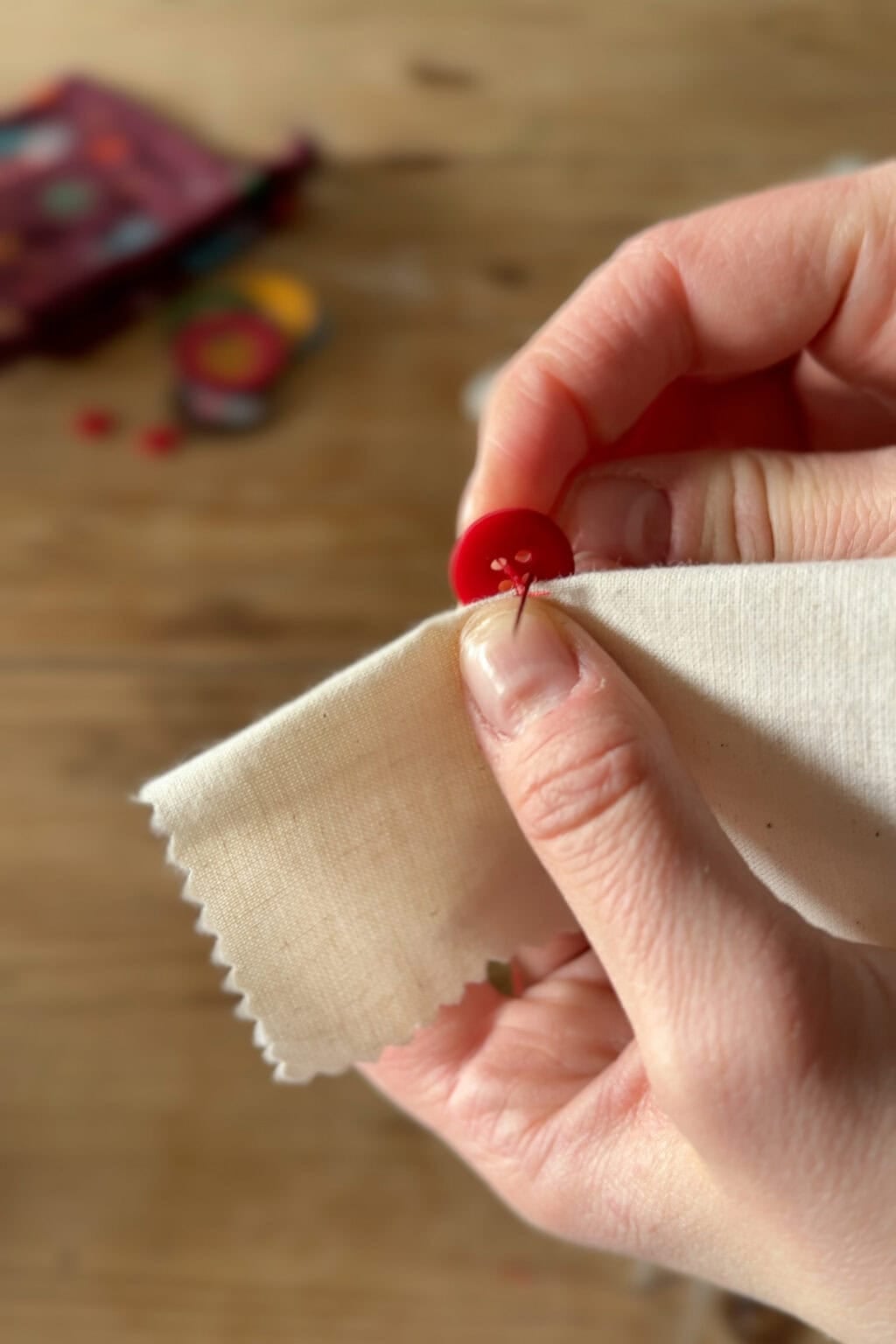 A close-up of hands sewing a red button onto a piece of off-white fabric with zigzag edges, using a needle and thread. The background is blurred with scattered sewing supplies.