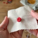 A person holds a piece of off-white fabric with pinked edges, displaying a red press stud attached to the cloth. The press stud is embossed with the word CHEEKY and a smiling face logo. The background is blurred.