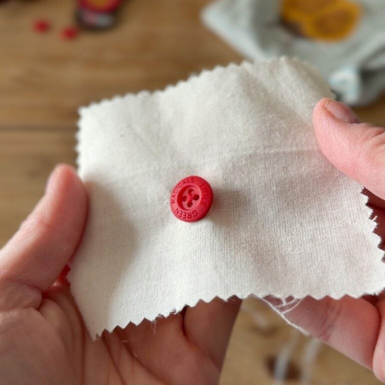 A person holds a piece of off-white fabric with pinked edges, displaying a red press stud attached to the cloth. The press stud is embossed with the word CHEEKY and a smiling face logo. The background is blurred.