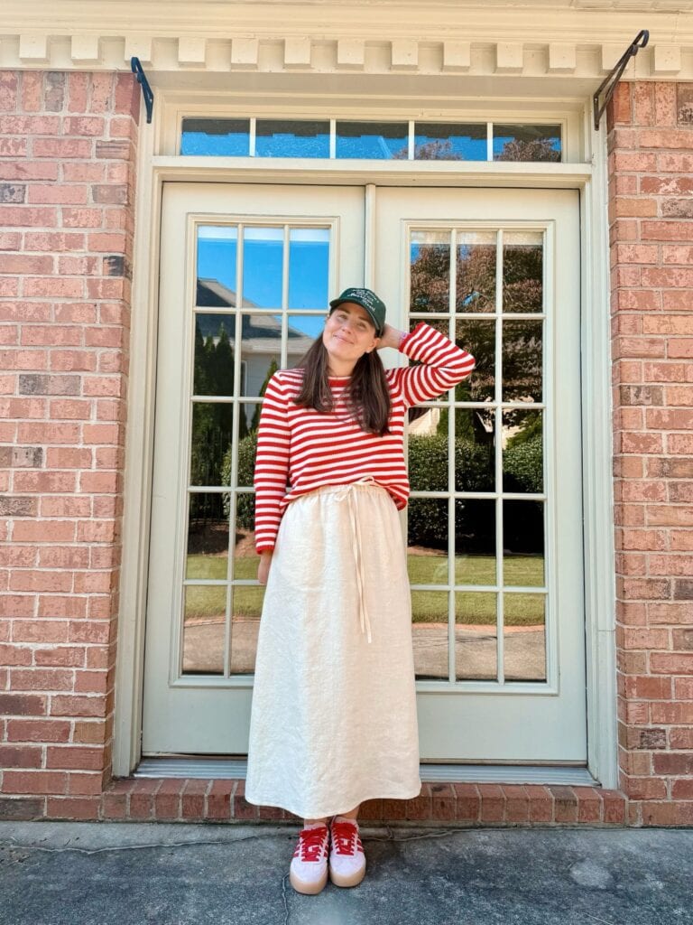 A person wearing a red-and-white striped long-sleeve shirt, a cream-coloured skirt (learn how to make a skirt out of a tablecloth!), green cap, and red trainers stands smiling in front of large glass doors with a brick wall background.