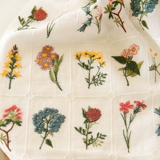A white cloth square featuring colourful, detailed embroidery of various wildflowers in a grid pattern—perfect inspiration if you’re learning how to make a skirt out of a tablecloth with unique floral designs.