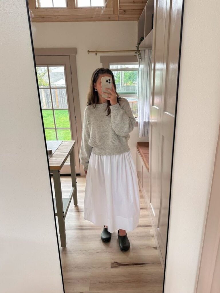 A person takes a mirror selfie in a bright room, wearing a grey jumper, black shoes, and a long white skirt styled like those from how to make a skirt out of a tablecloth tutorials. The space features wooden floors, large windows, and neutral-coloured walls.