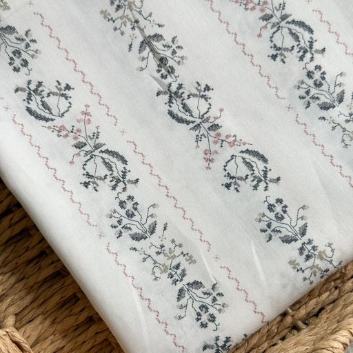 White fabric with a delicate, repeating floral pattern in grey and pink, laid on a woven straw surface—perfect inspiration if you’re wondering how to make a skirt out of a tablecloth in vintage style.