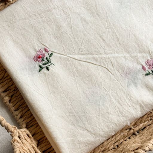 A cream-coloured fabric with small, delicate embroidered pink flowers and green leaves is draped over a woven basket, perfect inspiration if you’re wondering how to make a skirt out of a tablecloth.