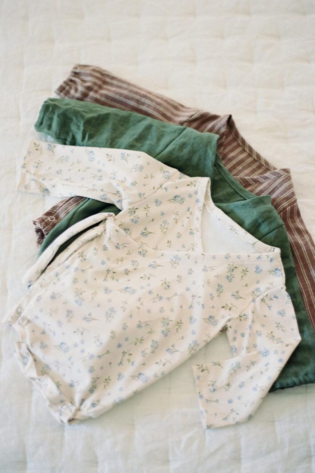 Three folded baby outfits are laid out on a light quilted surface: a cream-coloured babygrow with a small blue floral print, a green long-sleeved top, and a brown top with thin white stripes—adorable baby makes for beginner-friendly sewing.