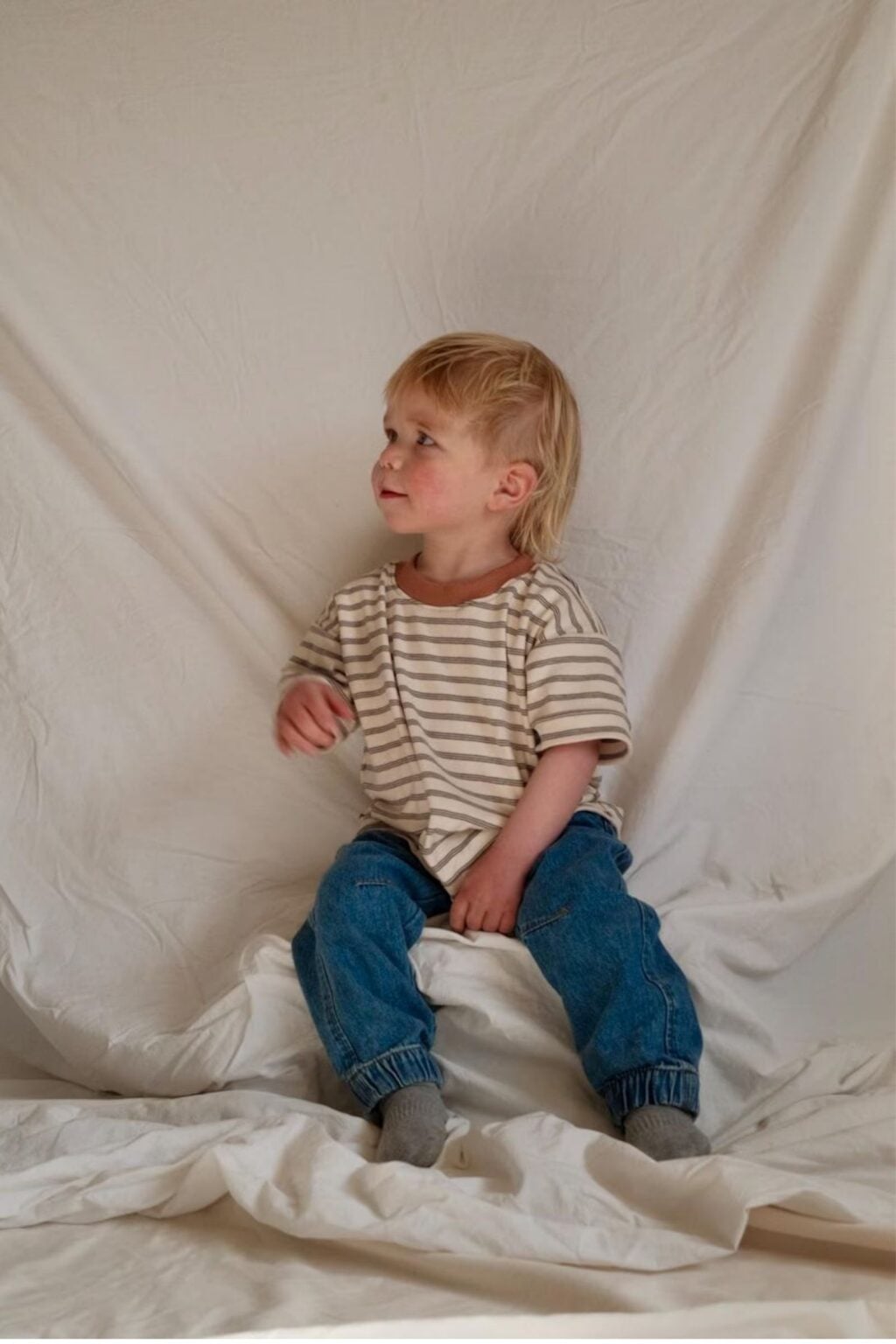 A young child with blonde hair, wearing a beige and white striped shirt and blue jeans, sits on a white cloth backdrop, looking to the left and smiling gently—perfect inspiration for adorable baby makes or beginner sewing projects.