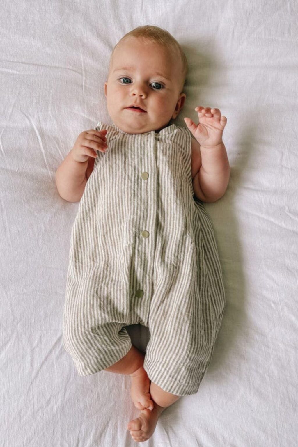 A baby wearing an adorable, sleeveless beige and white striped romper lies on a white sheet, looking up with one hand raised and legs slightly crossed—a perfect look for those seeking beginner-friendly baby sewing patterns.