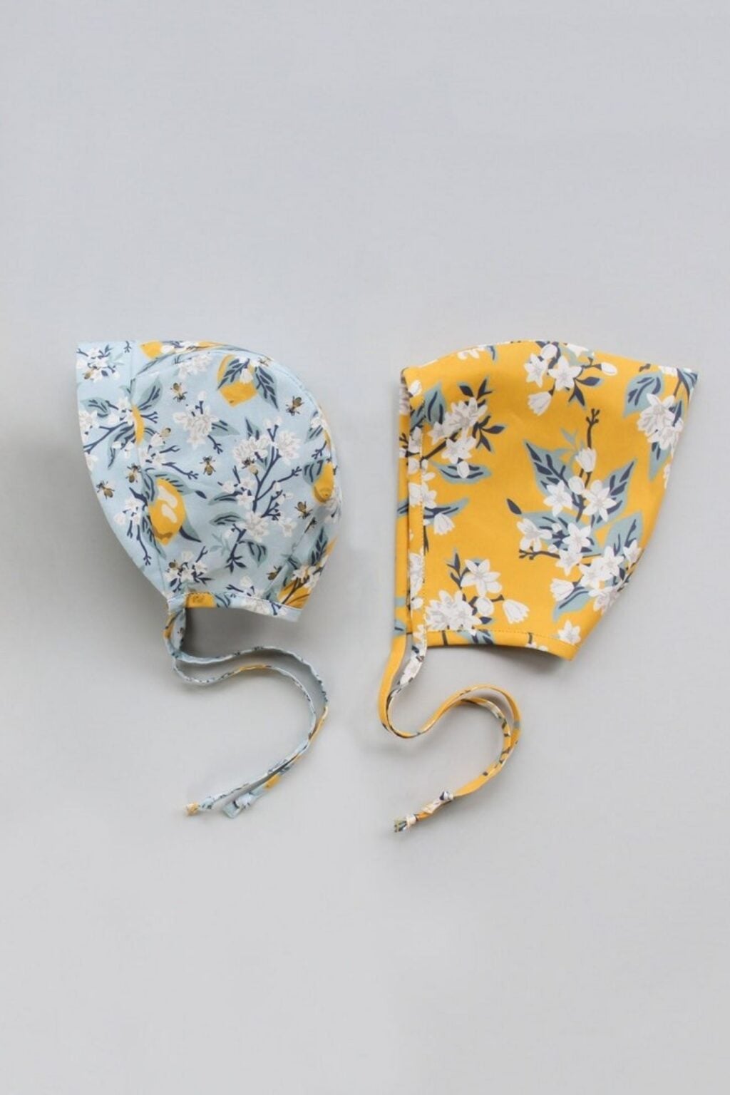 Two adorable bonnets are displayed on a light grey background: one is light blue with a lemon and floral pattern, the other is yellow with a white and blue floral print. Both have chin ties and are made using beginner-friendly baby sewing patterns.