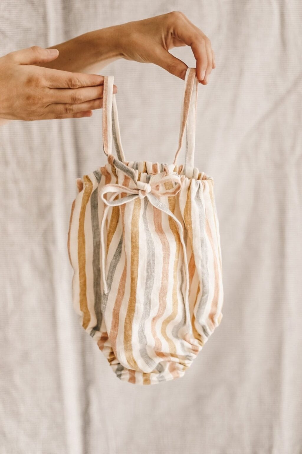 A hand holds up a striped baby romper with neutral tones of beige, mustard, and grey against a soft, textured fabric background—perfect inspiration for adorable baby makes and beginner-friendly sewing projects.