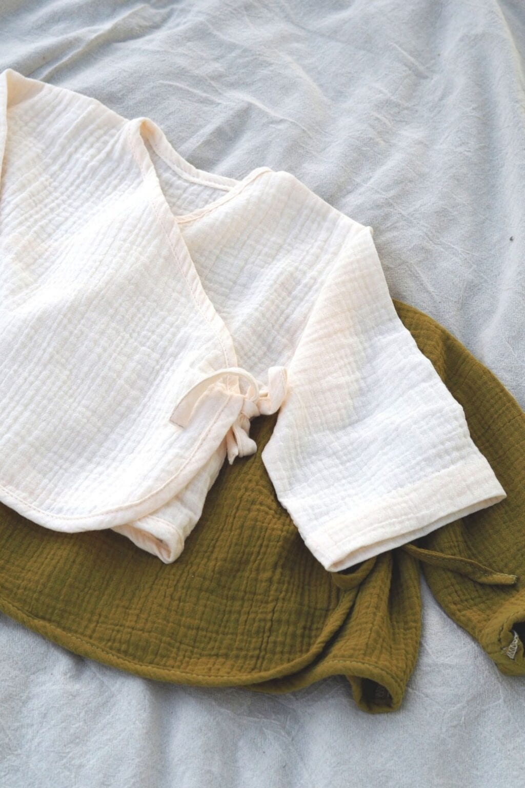Two baby kimono-style wrap tops, perfect for adorable baby makes or those using beginner-friendly sewing techniques, in cream and olive green muslin, are neatly folded on a light grey fabric background.