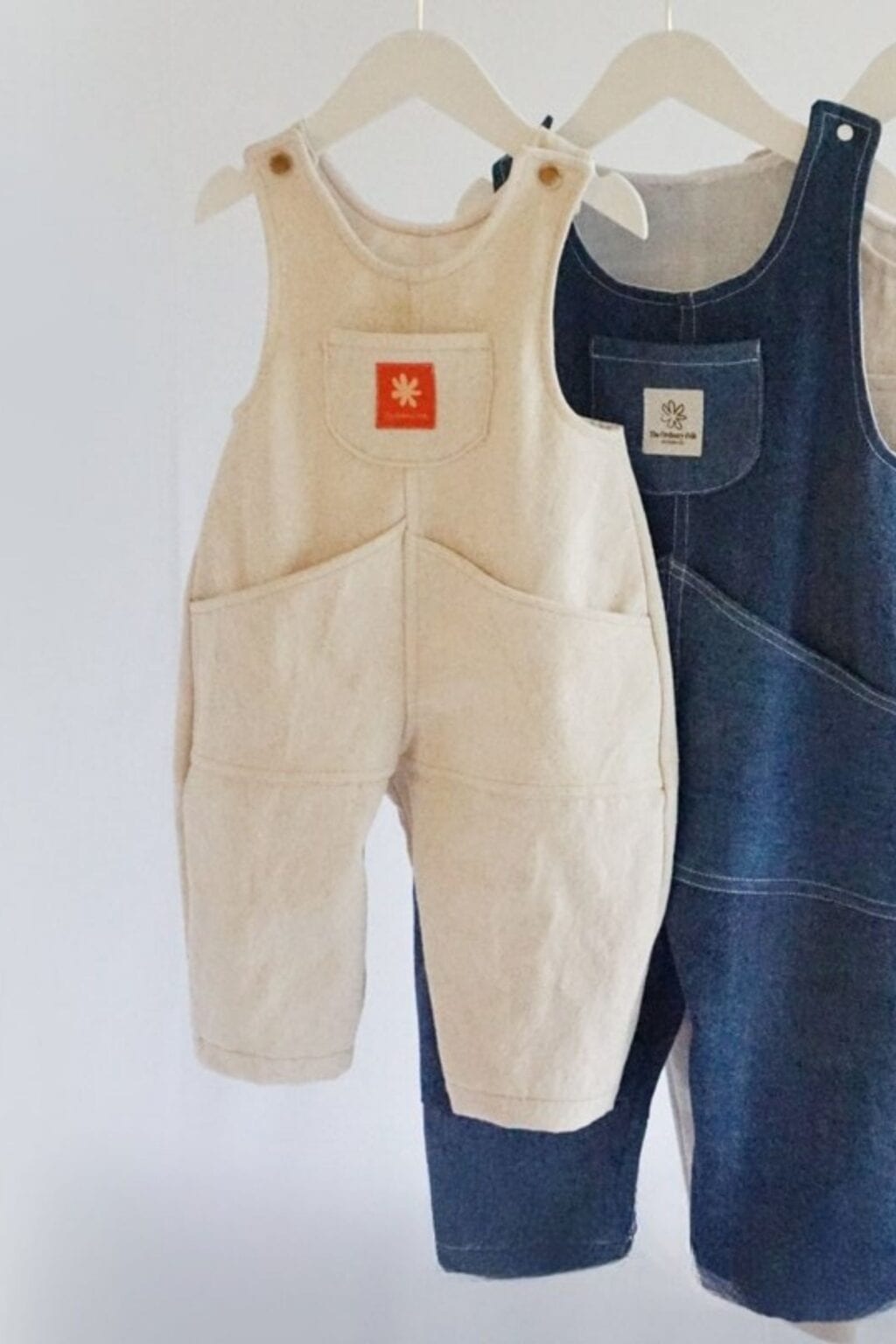 Two pairs of children's dungarees hanging on white hangers against a white background. Perfect for adorable baby makes, one pair is cream with an orange patch, and the other is dark blue denim with a white label.