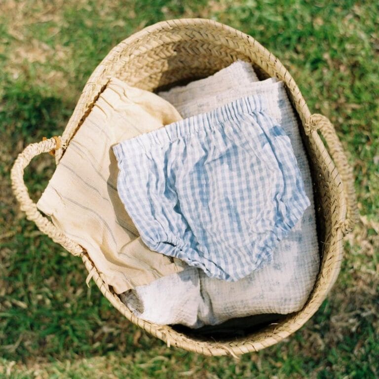 A woven basket sits on grass, holding folded light-coloured baby clothes, including a pair of blue and white gingham bloomers on top—perfect inspiration for adorable baby sewing and beginner sewing projects.