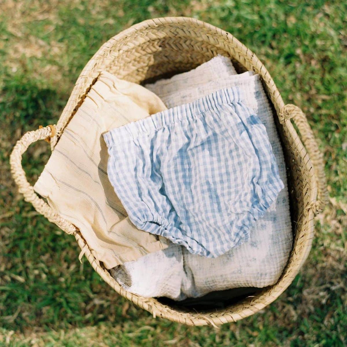 A woven basket sits on grass, holding folded light-coloured baby clothes, including a pair of blue and white gingham bloomers on top—perfect inspiration for adorable baby sewing and beginner sewing projects.